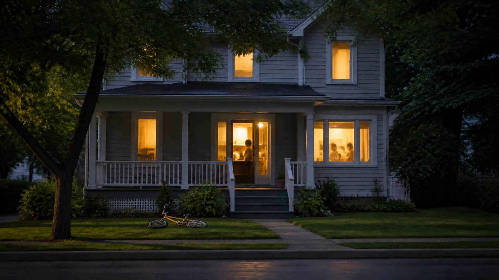 Family house at dusk with lit windows, soft silhouettes inside, and an amber child's bike on the lawn